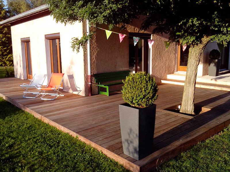 Aménagement extérieur avec une grande terrasse en bois, chaises longues colorées et banc vert devant une maisonnette en stuc, ensoleillée.