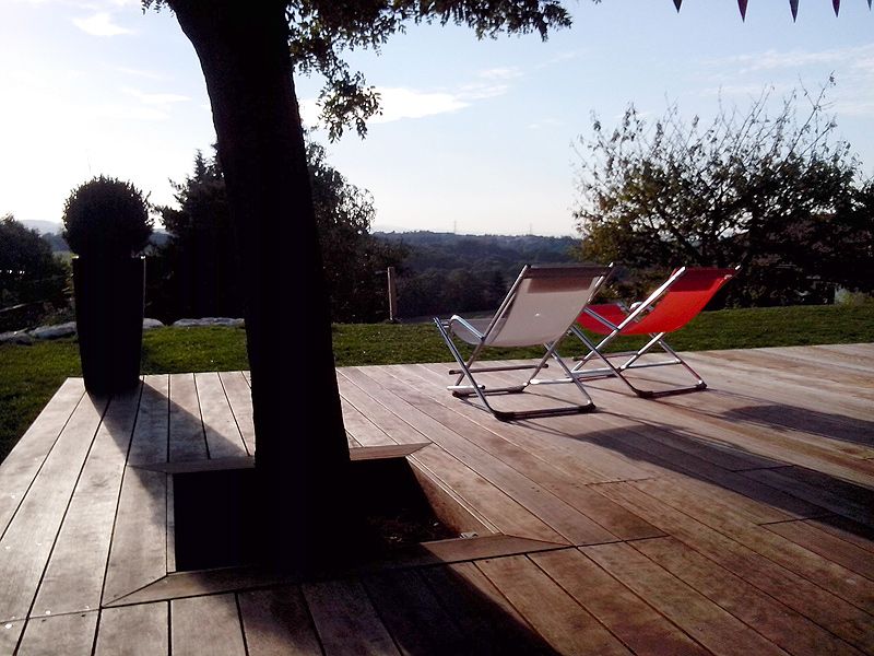Scène de détente sur une terrasse en bois intégrant un grand arbre. Deux chaises pliantes (rouge/blanc) et longues ombres.