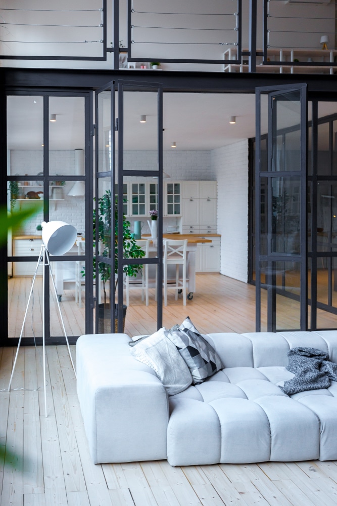 Verrière loft: séparation salon design et cuisine blanche. Salon moderne avec canapé gris et lampadaire, séparé d'une cuisine blanche lumineuse par une grande verrière industrielle ouverte.