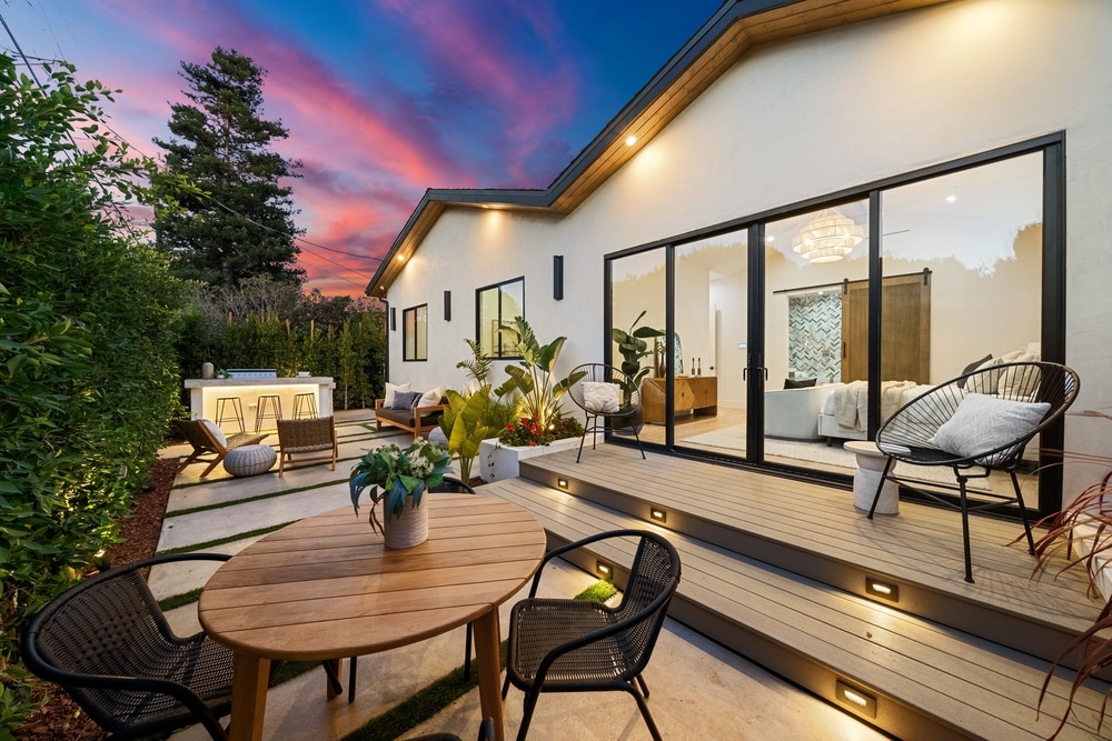 Patio et cuisine extérieure moderne au coucher du soleil Terrasse en bois et patio moderne au coucher du soleil, avec coin repas, cuisine extérieure illuminée et vue sur l'intérieur d'une chambre.