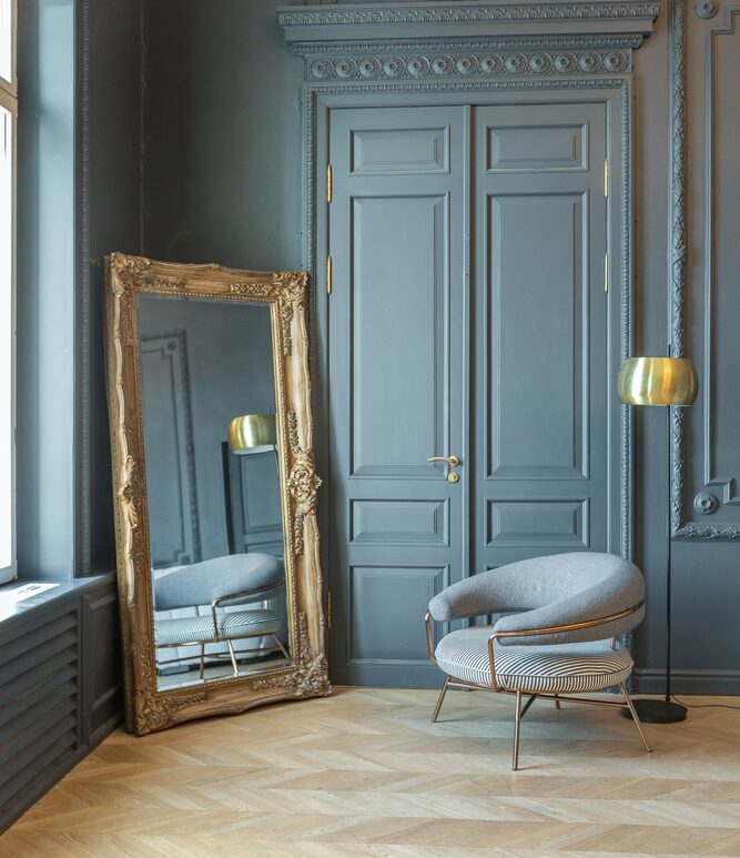 Mur bleu nuit mouluré, miroir doré et fauteuil design Intérieur bleu nuit: porte à moulures, miroir doré baroque et fauteuil design moderne. Parquet chevron en bois clair.