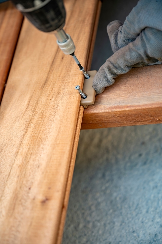 Fixation invisible : Installation facile d'une terrasse en bois. Installation de lames de terrasse en bois brun. Une main gantée visse un clip de fixation invisible avec une perceuse.