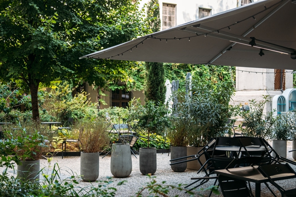 Terrasse Jardin Ombragée : Design et Ambiance Végétale Terrasse jardin ombragée, grand parasol avec lumières, mobilier empilé, pots en béton et végétation luxuriante.